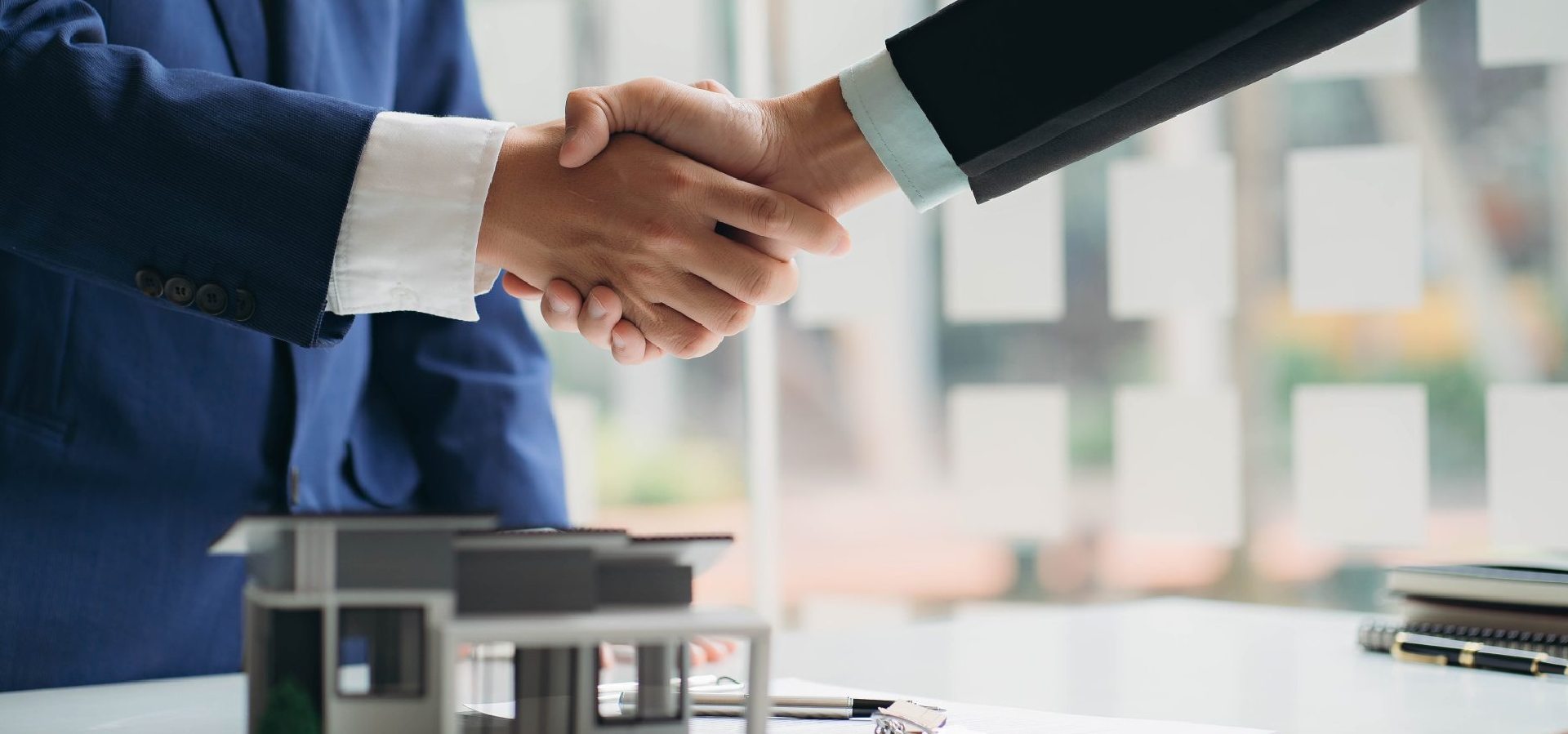 Dos personas de traje se dan la mano sobre la miniatura de una casa